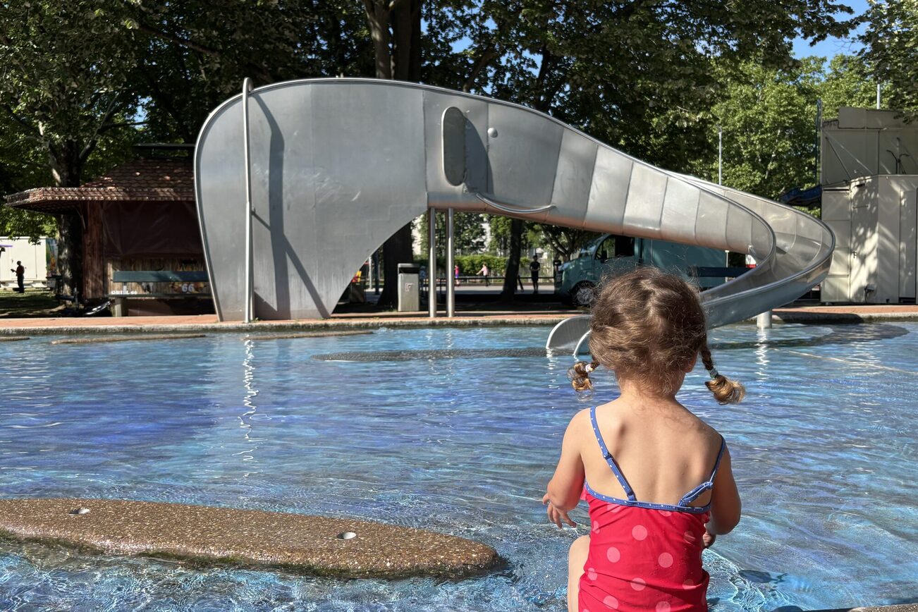 Dziewczynka w czerwonym kostiumie kąpielowym w kropki siedzi przy basenie i patrzy w kierunku zjeżdżalni w kształcie słonia.