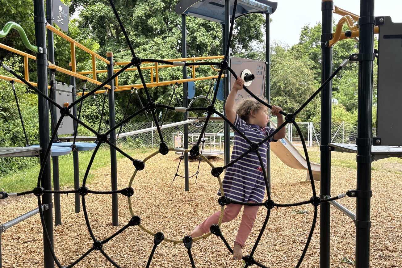 Plac zabaw dla dzieci z drabinką, dziewczynka w fioletowo-białej sukience w paski i różowych spodniach wspina się.
