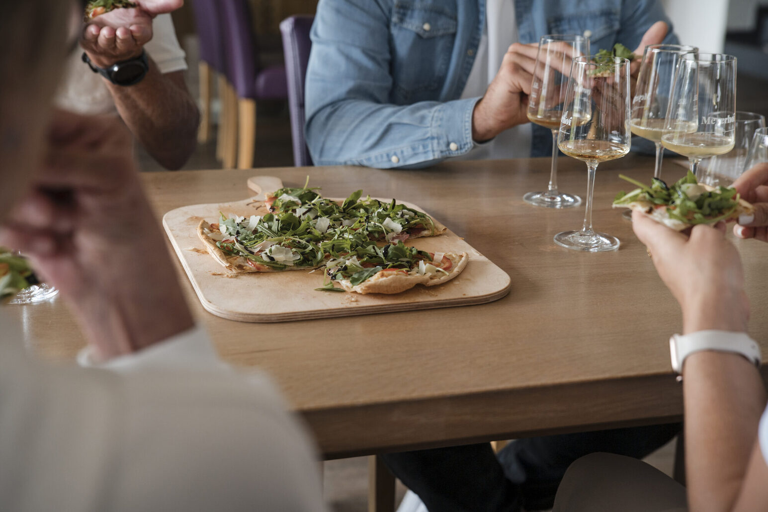 Una mesa con tarta flambeada y manos que sostienen trozos de tarta flambeada. En la mesa hay cuatro copas de vino blanco.