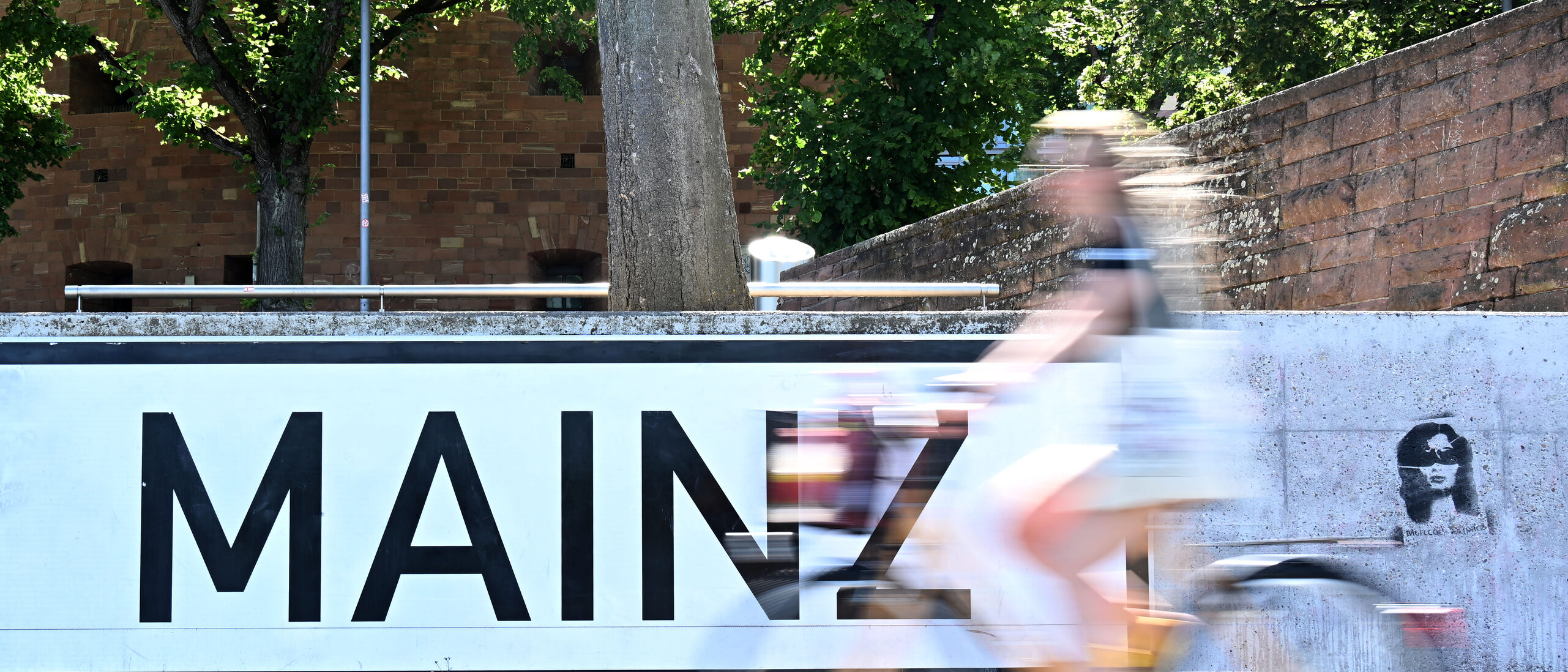 Napis MAINZ wielkimi literami, czarno na białym na szarej ścianie, przed nim rowerzysta (niewyraźny)