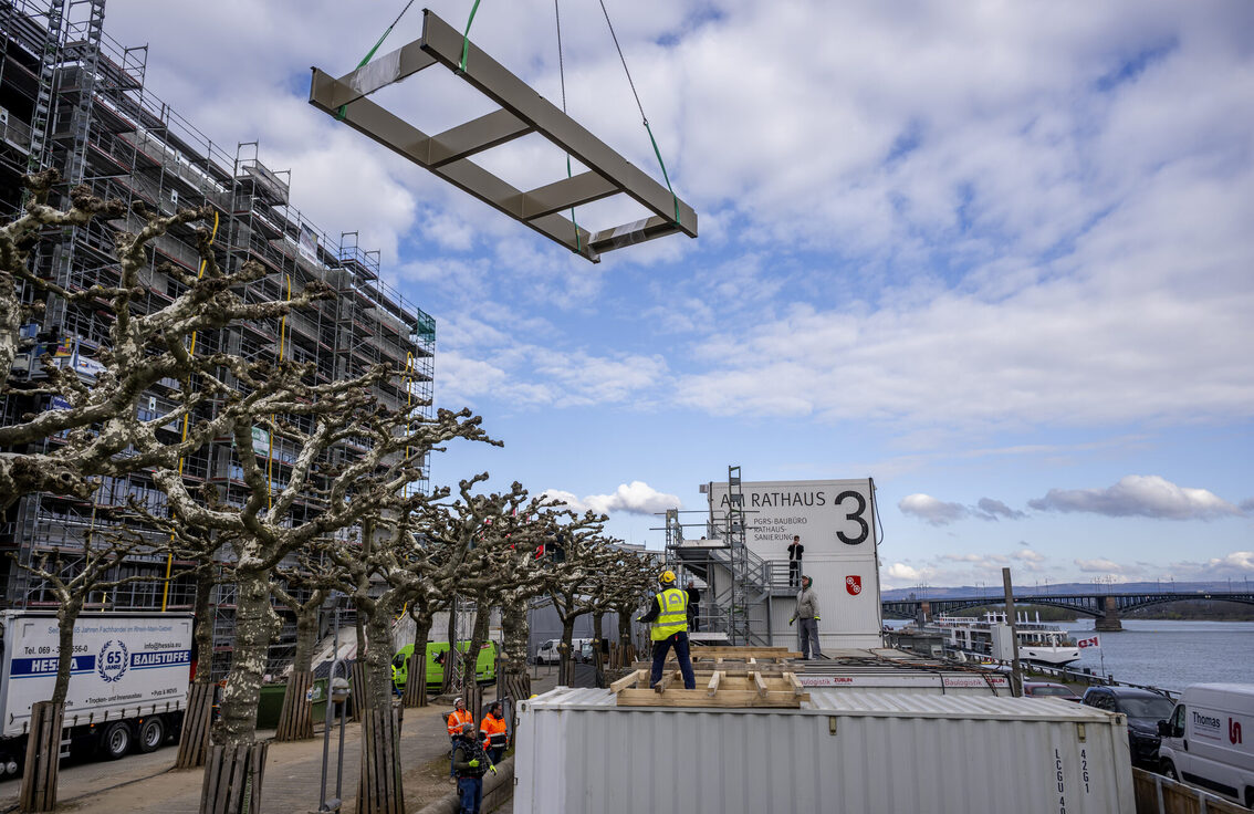 Ren Nehri kıyısında bir konteynerin üzerinde duran inşaat işçisi ve üzerinde bir vinç üzerinde asılı duran bir bileşen