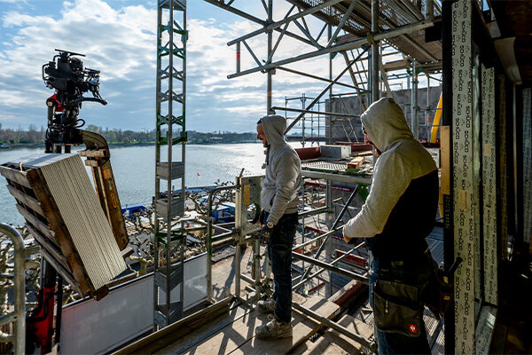 İki inşaat işçisi iskelenin üzerinde duruyor, solda yüklü vinç, arka planda Ren Nehri.