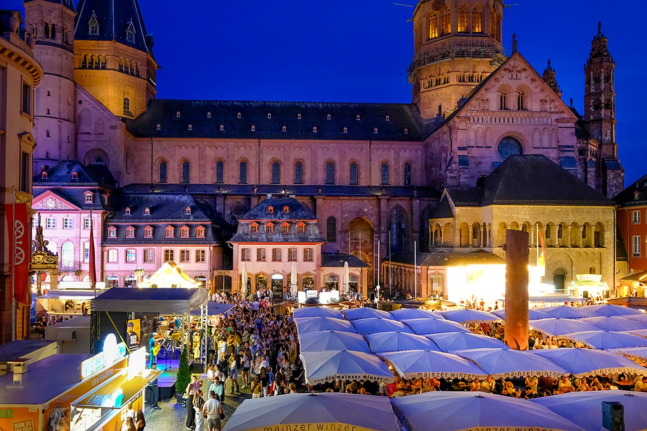 Blick von oben auf den Markt, der gefüllt ist mit Menschen, die unter weißen Schirmen sitzen. Links steht eine Bühne, im Hintergrund ist der Dom.