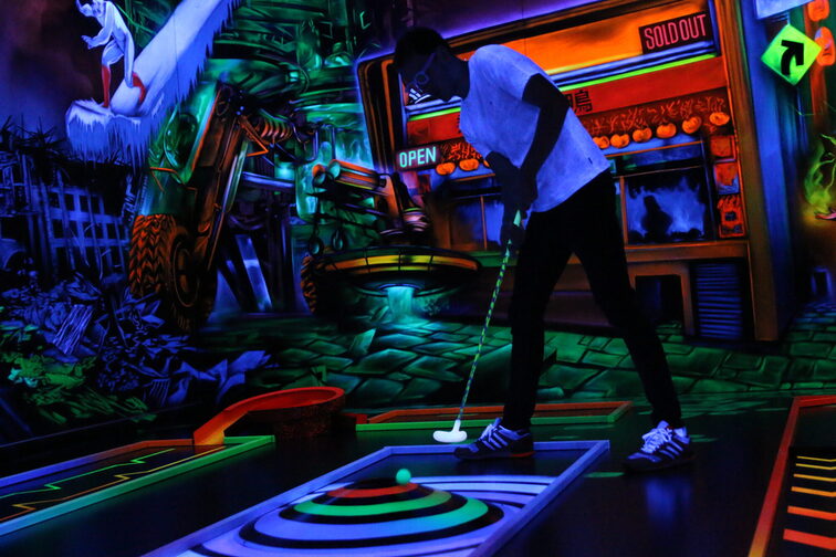 Un joueur de minigolf sur une piste en lumière noire devant des graffitis au néon