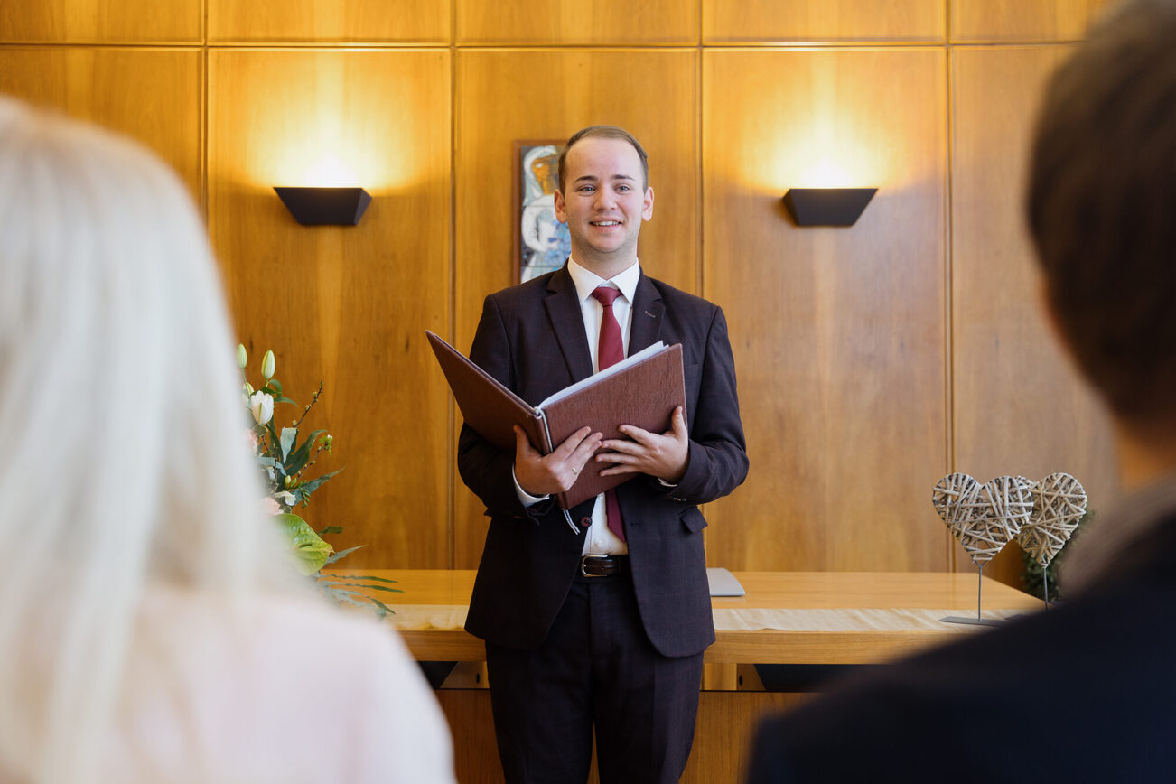 يقف أمين السجل في المنتصف ومعه كتاب بني اللون مفتوح، ويمكن رؤية مؤخرة رأس امرأة ورجل في المقدمة يميناً ويساراً.