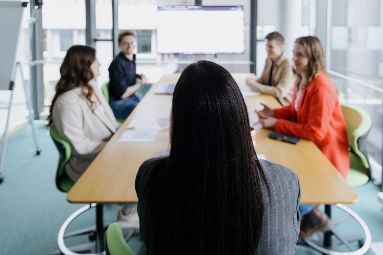 5 junge Mitarbeiter:innen sitzen an einem Tisch, im Hintergrund ein Bildschirm.